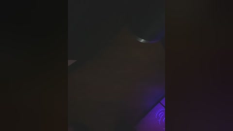 Media: A dimly lit, close-up video of a woman's face with dark skin, partially illuminated by purple light. Her facial expression is indiscernible due to the low lighting. The background is dark and indistinct, suggesting an intimate or private setting.