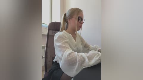 Media: Video of a blonde woman with glasses, wearing a sheer white blouse, sitting on a brown chair in a bright, minimalistic room with a window.