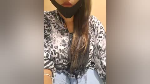 Media: Video of a woman with long, straight brown hair, wearing a black blindfold, leopard-print blouse, and light blue jeans, sitting indoors.