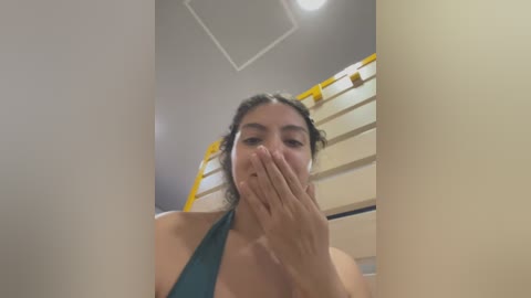 Media: Video of a woman with light brown skin and dark hair partially covering her face, wearing a teal halter top, standing in a stairwell with beige walls and yellow railings.