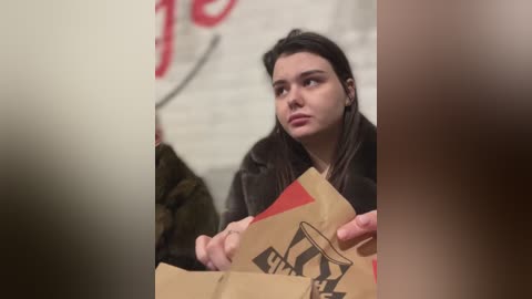 Media: Video of a young woman with long brown hair, wearing a dark coat, holding a brown paper bag with a red and black logo. Background features blurred text and a white brick wall.