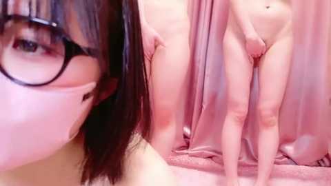 Media: Video of a fair-skinned woman with dark hair and glasses, covering her mouth with her hand, standing behind a pink curtain. Her nude body is partially visible, holding her thighs.