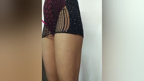 Media: Video of a woman's lower body in a black, polka-dot mini dress with a sheer, fishnet panel and a red lace overlay. Her skin is light brown, and she stands against a plain white wall.