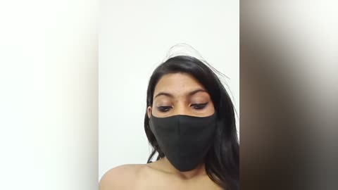 Media: A video of a young woman with medium skin tone, wearing a black face mask, black bra, and long black hair. She stands against a plain white background with a blurred brown wall on the right.