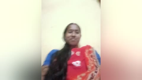 Media: A blurry video of a South Asian woman with medium brown skin and long black hair, wearing a red sari with white floral designs, standing against a pale yellow wall.