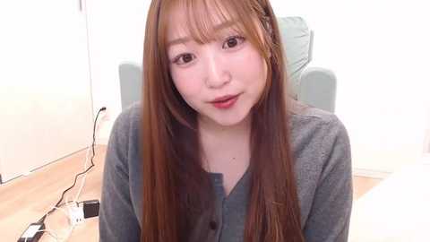 Media: A video of an Asian woman with long, straight, auburn hair, fair skin, and brown eyes, wearing a gray cardigan, sitting in a light-colored office chair against a white wall.