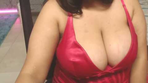 Media: A close-up video of a woman with fair skin, wearing a red satin camisole with a deep V-neck, revealing ample cleavage. She sits indoors near a pool with a tiled floor.