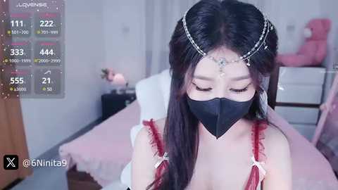 Media: Video of an East Asian woman with long black hair, wearing a black face mask, red lingerie, and a tiara, in a bedroom setting with a pink bed and dresser.