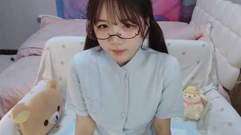 Media: Video of an East Asian woman with glasses, light skin, and brown hair in pigtails, wearing a light blue button-up shirt. She sits on a white bed with pink pillows and stuffed animals, including a teddy bear and a rabbit.