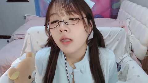 Media: A video of an Asian woman with long black hair in pigtails, wearing glasses and a light blue blouse, lying on a pink bed, surrounded by colorful pillows.