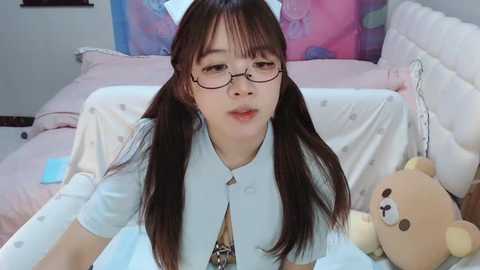 Media: Video of an East Asian woman with long brown hair and glasses, wearing a white blouse and tie, lying on a bed with pink and white bedding, holding a teddy bear.