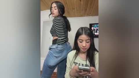 Media: Video of a young woman with long black hair, wearing a striped crop top and baggy jeans, looking over her shoulder. Another woman with long black hair, wearing a white T-shirt, is focused on her phone.