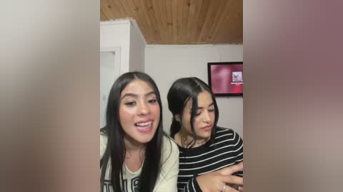 Media: Video of two young women, one with long black hair and light skin, smiling, the other with dark hair, wearing a striped shirt, indoors with wooden ceiling and framed picture.