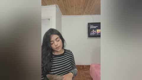 Media: Video of a young woman with long black hair, wearing a black and white striped shirt, sitting on a pink couch in a small, cozy room with wooden ceiling and a TV mounted on the wall.