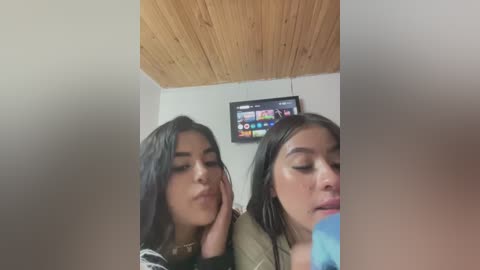 Media: Video of two young women with long black hair, making kissing faces while sitting in a wooden-paneled room with a TV showing a colorful channel guide.