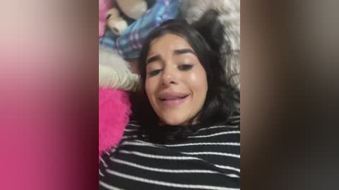 Media: Video of a young woman with long dark hair, lying on her back in a bed, surrounded by colorful stuffed animals. She wears a black and white striped shirt, and her face is relaxed and slightly smiling.