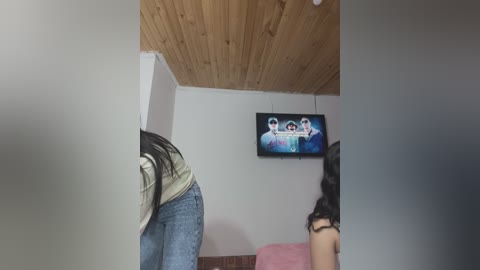 Media: Video of a woman with long black hair bending over in tight jeans, standing in a room with wooden ceiling, pink couch, and TV showing three men in a sports jersey.