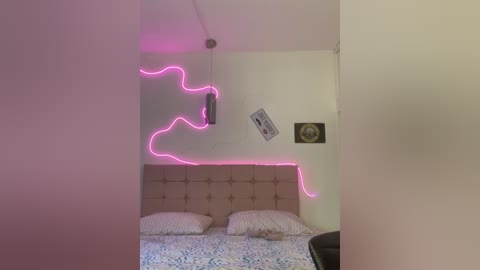 Media: Video of a minimalist bedroom with a tufted beige headboard, two pillows, a blue and white floral duvet, and a neon pink zigzag light strip on the wall.