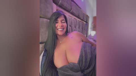 Media: A video of a smiling Latina woman with long, dark hair, large breasts, and a dark grey towel covering her top half, sitting against a padded grey wall in a dimly lit room.