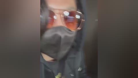 Media: A blurry video of a person wearing a black hoodie and reflective sunglasses, standing indoors with a blurred background.