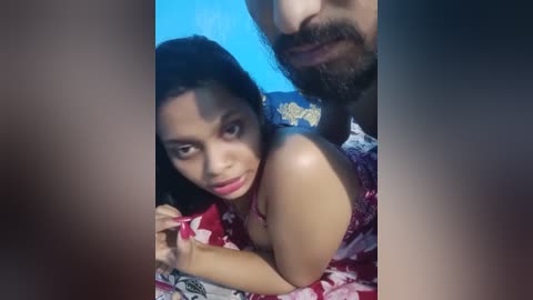 Media: Video of a young woman with medium skin tone and dark hair, wearing a red floral top, sitting on a bed. A bearded man with a mustache stands behind her, holding her shoulders.