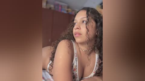 Media: Video of a young woman with curly hair, wearing a floral tank top, leaning forward in a bedroom with a blurred background of shelves filled with books and items.