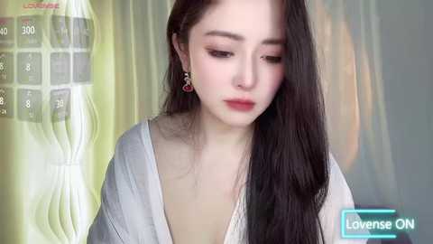 Media: Video of a young woman with long, straight dark hair, fair skin, and red lipstick, wearing a white blouse, looking contemplative. Background features a blurred, pastel-colored wall and a calendar.