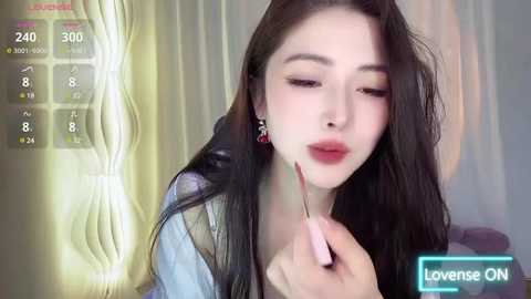 Media: Video of a young Asian woman with fair skin, long black hair, and makeup, applying lipstick in a bedroom setting with soft lighting.