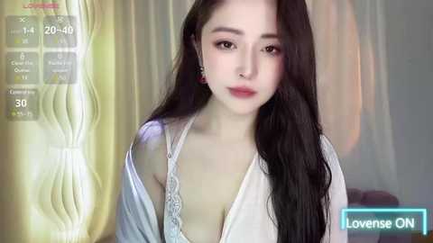 Media: Video of a young East Asian woman with long, straight black hair, wearing a white lace bralette, standing in a softly lit room with cream curtains.