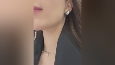 Media: Video of a woman's face and upper chest, focusing on her lips, fair skin, and a black blazer with a silver necklace and diamond earrings. The background is out of focus.