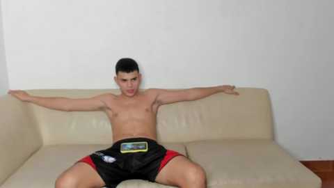 Media: Video of a shirtless young man with short black hair, wearing black shorts with red trim, lying on a beige leather couch with arms stretched out. The background is a plain white wall and a wooden floor.