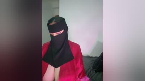 Media: A video of a person wearing a black niqab with a red garment underneath, standing in a dimly lit room with white walls.