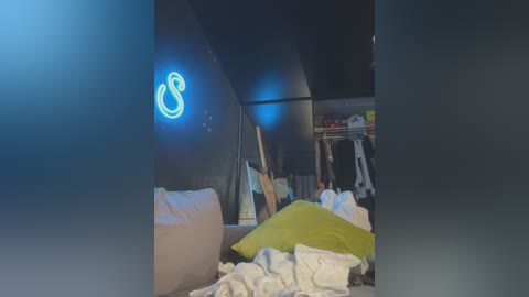 Media: Video of a messy bedroom with neon blue \"B\" sign, white and green pillows, clothes scattered on the floor, and a closet visible in the background.