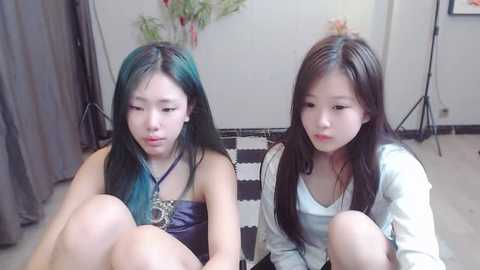 Media: Video of two young East Asian women with long, straight black hair, wearing casual outfits, sitting on a patterned rug in a minimalist room with a floral arrangement in the background.