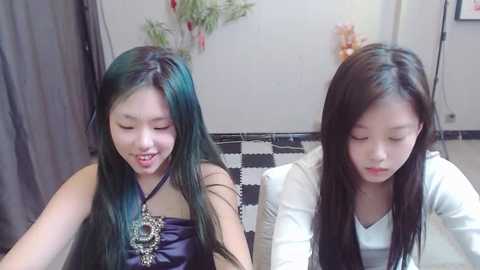 Media: Video of two Asian girls with long black hair, one wearing a dark dress, the other a white top, sitting on a black-and-white tiled floor, smiling.