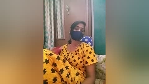 Media: A video shows an Indian woman in a yellow sari with black floral patterns, wearing a face mask, sitting on a bed with blue polka dot sheets, in a modest room with green and white curtains.