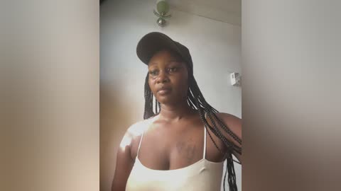 Media: Video of a dark-skinned woman with braided hair, wearing a black baseball cap and white tank top, standing in a dimly lit room with white walls.