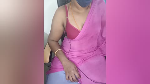 Media: Video of a woman with medium brown skin, wearing a pink saree, a red bra, and blue pants, seated on a wooden chair, with a gray mask covering her nose and mouth.