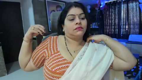 Media: Video of a South Asian woman in a patterned orange blouse, holding a white sari, with a serene expression. Background includes a framed picture and dark curtains.