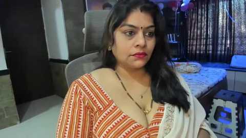 Media: Video of a South Asian woman with long black hair, wearing a traditional orange and white saree, seated indoors, with a modern room in the background.