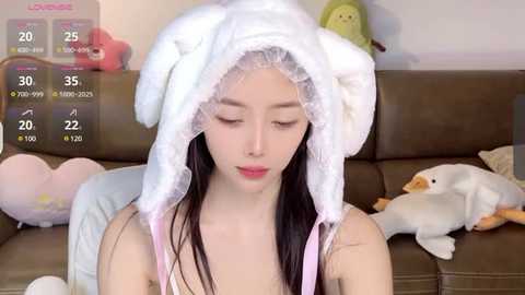 Media: Video of a young Asian woman with fair skin, wearing a fluffy, white bunny-themed hood and pink lingerie, sitting on a brown couch, surrounded by plush toys.