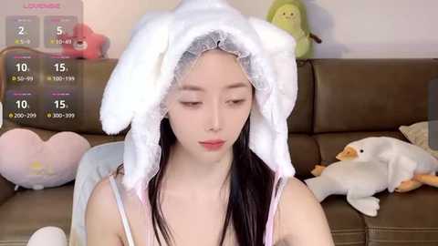 Media: Video of an East Asian woman with long black hair, wearing a white, plush cat hat, seated on a brown couch, surrounded by plush toys, including a duck and a heart-shaped pillow.