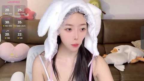 Media: Video of an East Asian woman with fair skin, long black hair, and a white cat hat, sitting on a brown leather couch. The background features stuffed animals and a digital weather display.