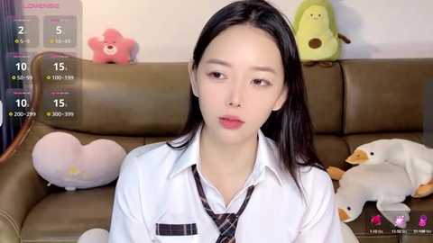 Media: Video of a young East Asian woman with fair skin and straight black hair, wearing a white shirt and black tie, seated on a brown leather couch with plush toys, including a pink star, a yellow frog, and a white duck.