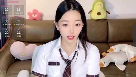 Media: Video of an Asian woman with long black hair, fair skin, wearing a white shirt and tie, sitting on a brown couch with plush toys.