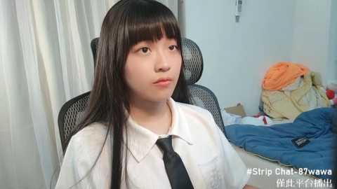 Media: Video of a young Asian woman with long black hair, wearing a white shirt and black tie, sitting in a black mesh chair. Behind her, a messy bed with orange and blue sheets.