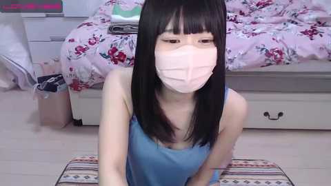 Media: Video of an Asian woman with straight black hair and pale skin, wearing a blue tank top and a pink surgical mask, kneeling on a striped rug in a bedroom with a floral bedspread and white dresser.