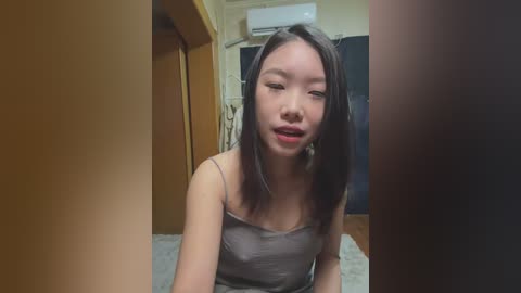 Media: Video of a young East Asian woman with straight black hair, wearing a spaghetti-strap gray top, sitting on a bed in a modestly furnished room with wooden furniture and a window air conditioner.