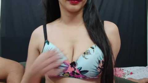 Media: Video of a light-skinned woman with long black hair, wearing a floral-patterned bra with pink and purple flowers, adjusting the straps. Background shows a dark curtain and a bed with white sheets.