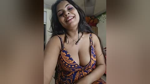 Media: Video of a smiling South Asian woman with medium skin tone, wearing a low-cut, patterned dress, indoors.